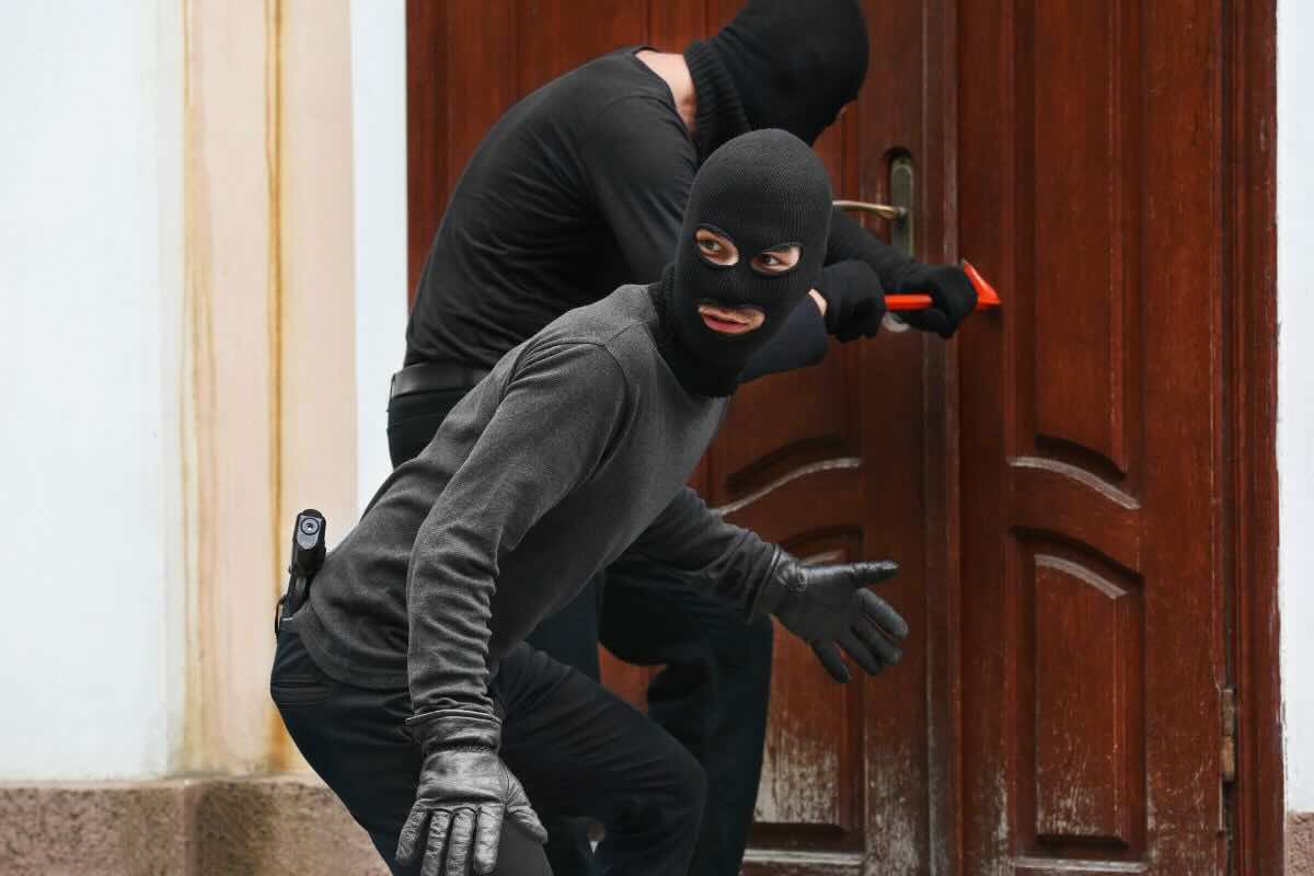Due uomini vestiti di nero con passamontagna: uno fa da guardia e l'altro cerca di scassinare porta in legno marrone con un arnese rosso