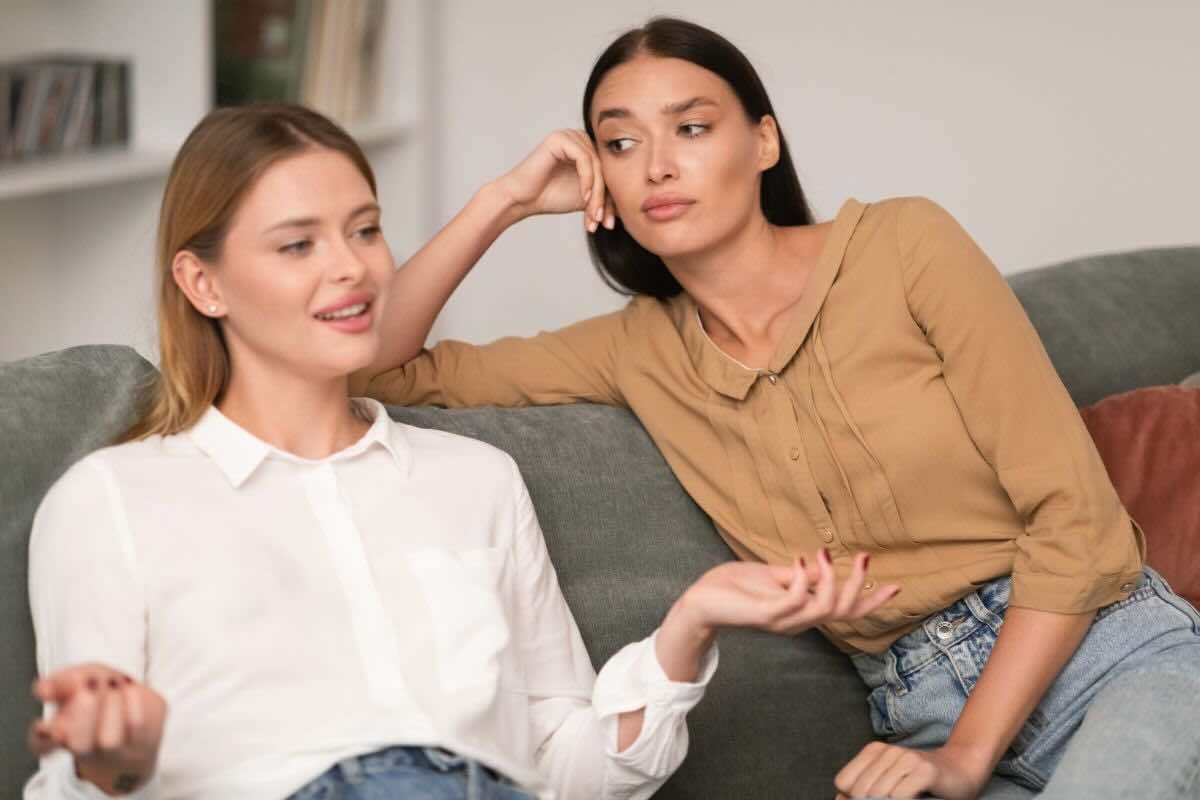 Donne sedute sul divano: una parla sorridente, l'altra la guarda invidiosa