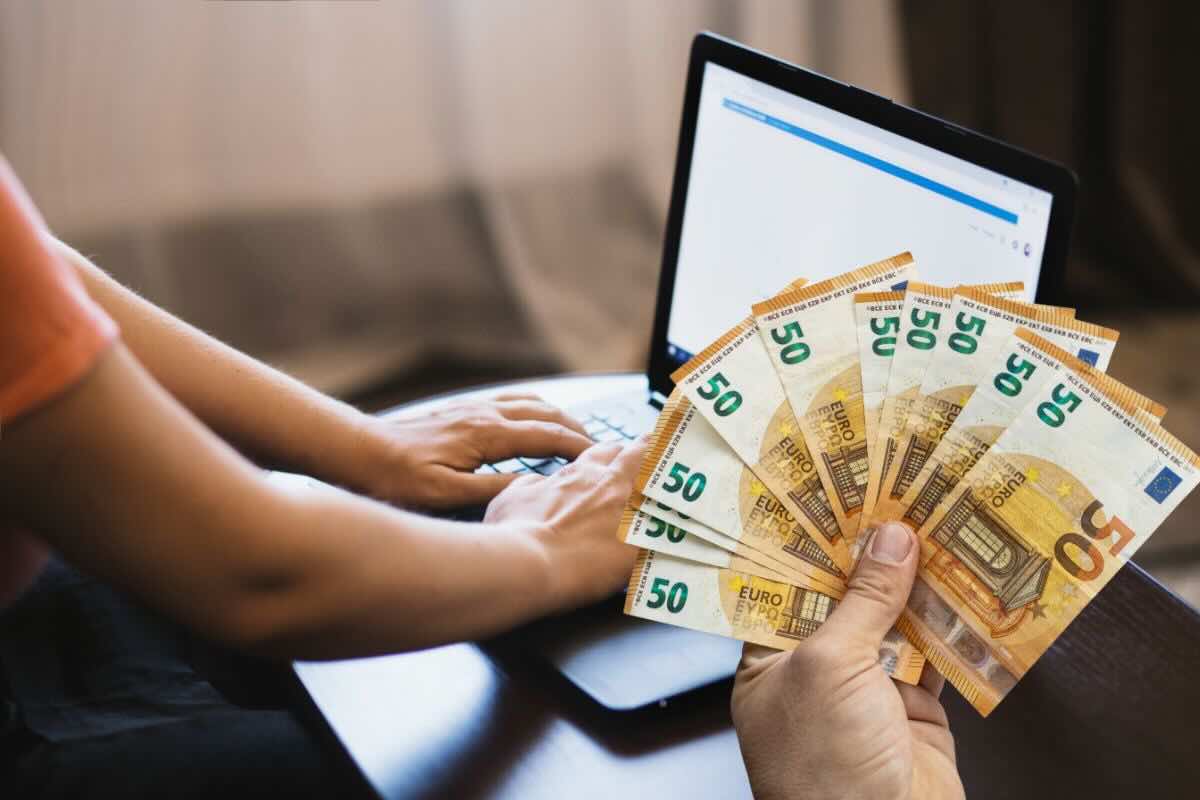 Persona scrive su tastiera PC, in primo piano mano tiene banconote da 50 euro