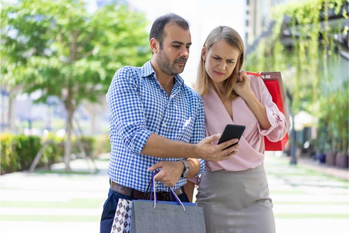 Uomo e donna buste shopping, lui con cellulare in mano guarda schermo, lui viso corrucciato
