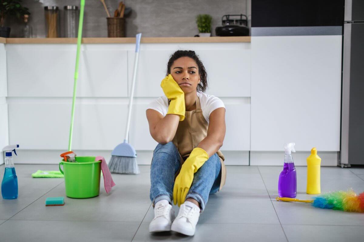 Ragazza seduta sul pavimento un po' annoiata, indossa guanti gialli per le pulizie. Intorno a lei strumenti e prodotti per fare le faccende domestiche