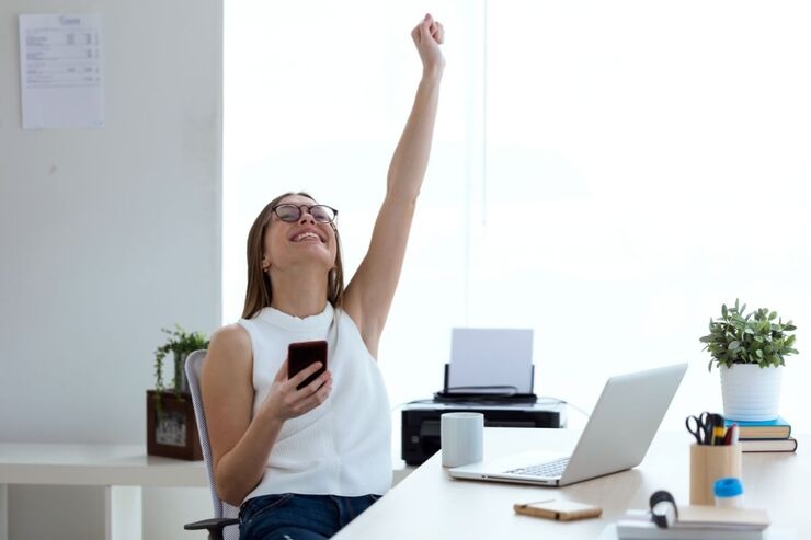 Donna esulta con smartphone in mano, seduta alla scrivania davanti al PC